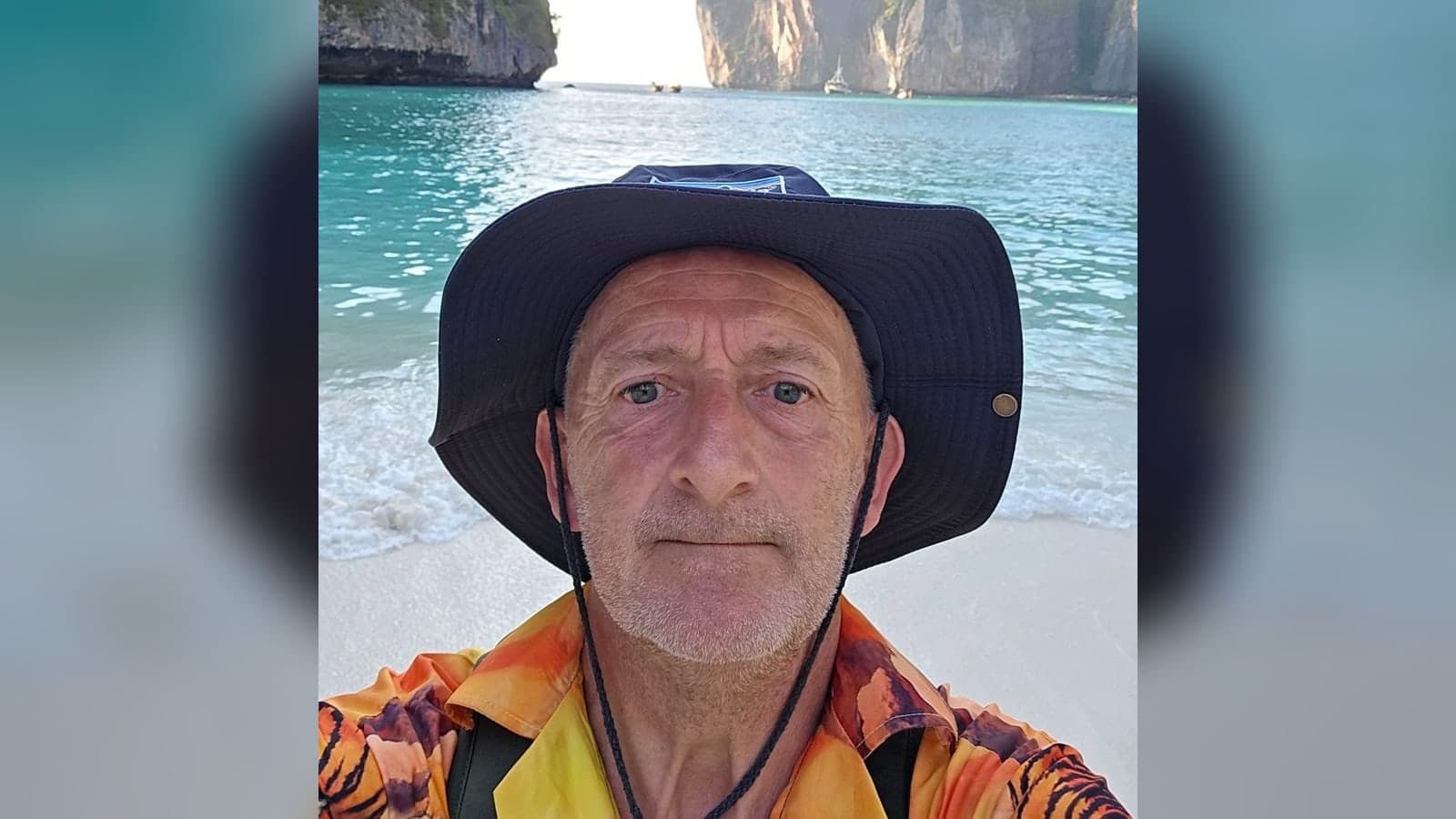 Donny Knight in a navy wide-brimmed hat and an orange tiger-print shirt, taking a selfie on a beach